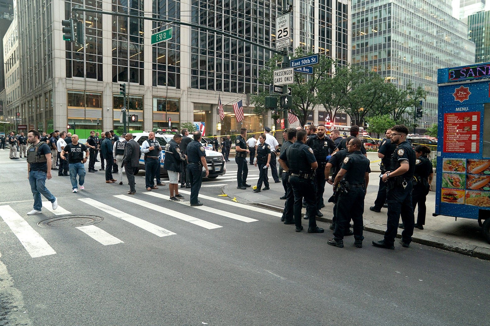 A taped-off crime scene area on the street is surrounded by clusters of bystanders and lots of police officers.