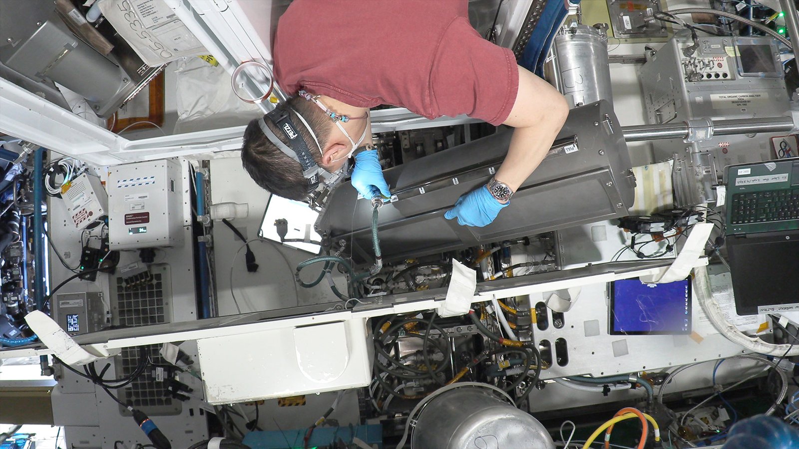 a man in a maroon shirt and safety goggles is seen from behind working to install a tank aboard a space station