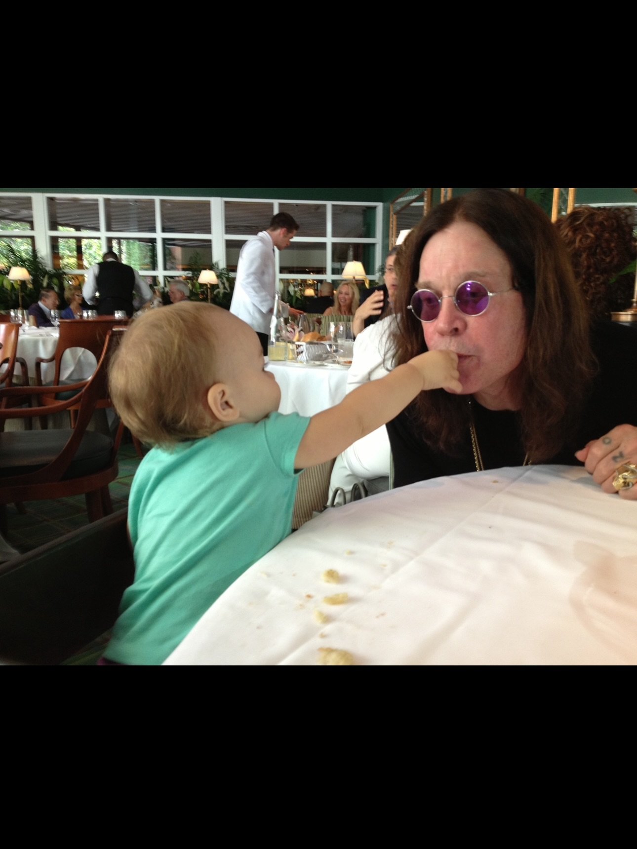 Ozzy Osbourne is fed by an infant at a table. 