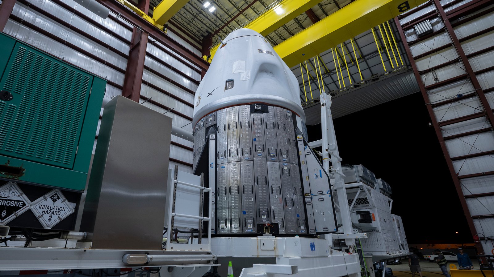 a conical white space capsule inside a hangar, with the night sky visible through a large open door behind it