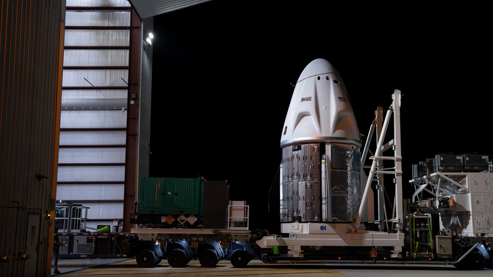 a white conical space capsule is wheeled into a hangar at night