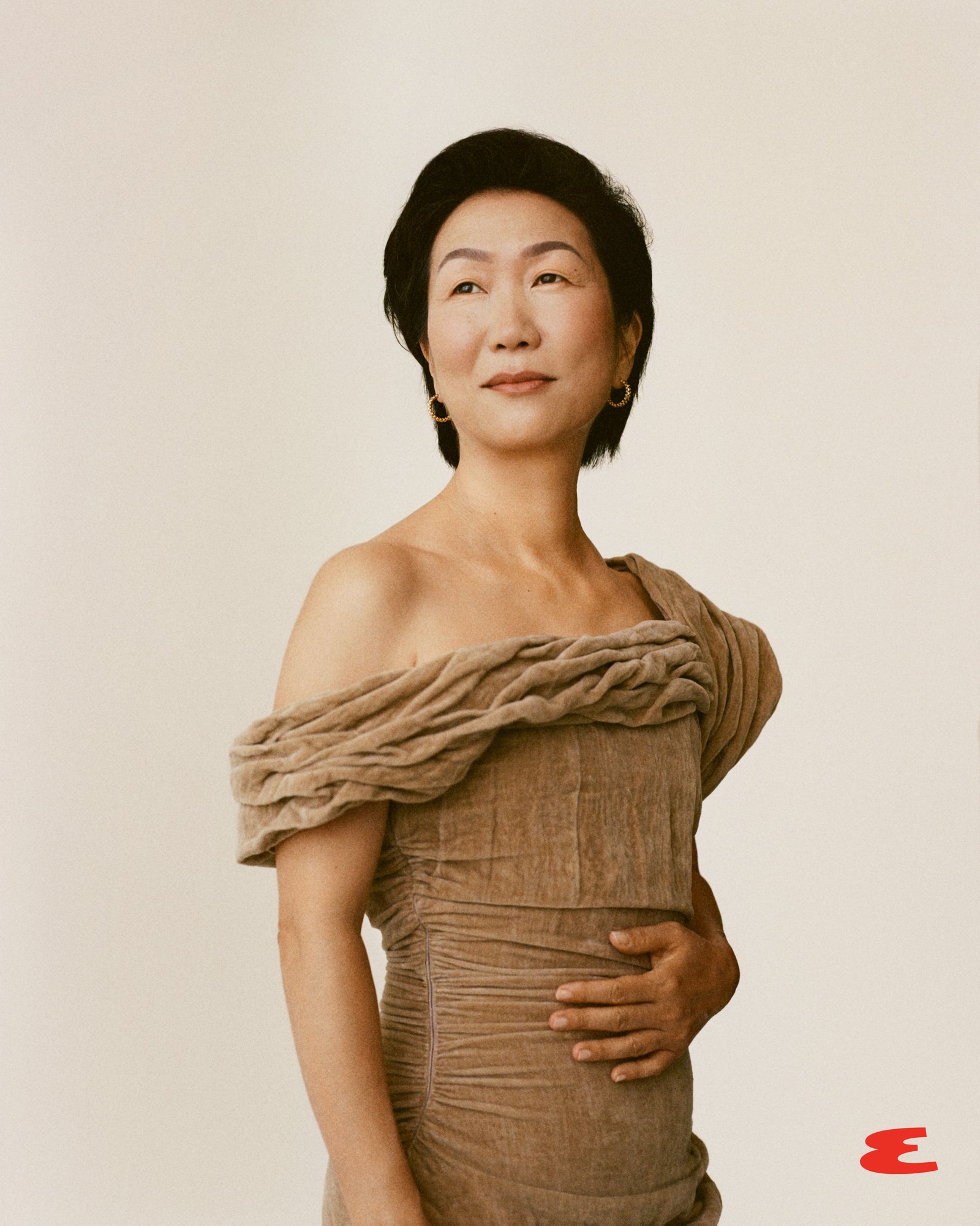 a figure dressed in a uniquely styled brown garment poses with one hand on their midsection