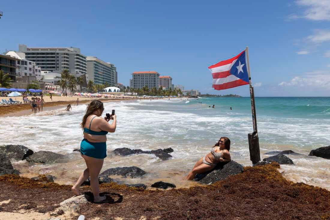 A woman poses for photos next to a Puerto Rican flag along the beach in Condado, Puerto Rico, as Hurricane Erin approaches, Friday, Aug. 15, 2025.