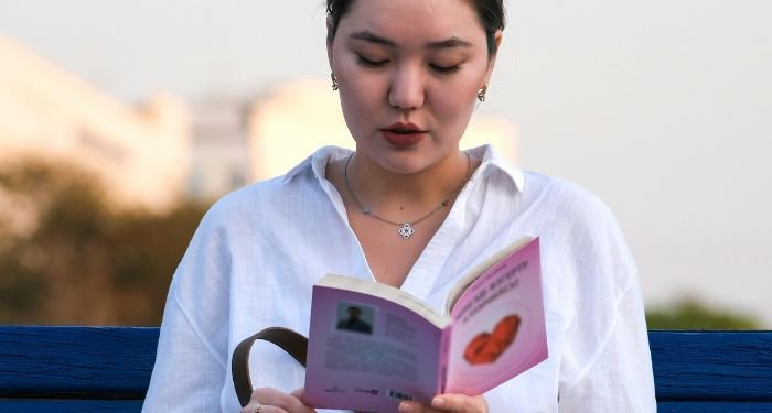 fair-skinned dark haired woman reading a book on a blue bench
