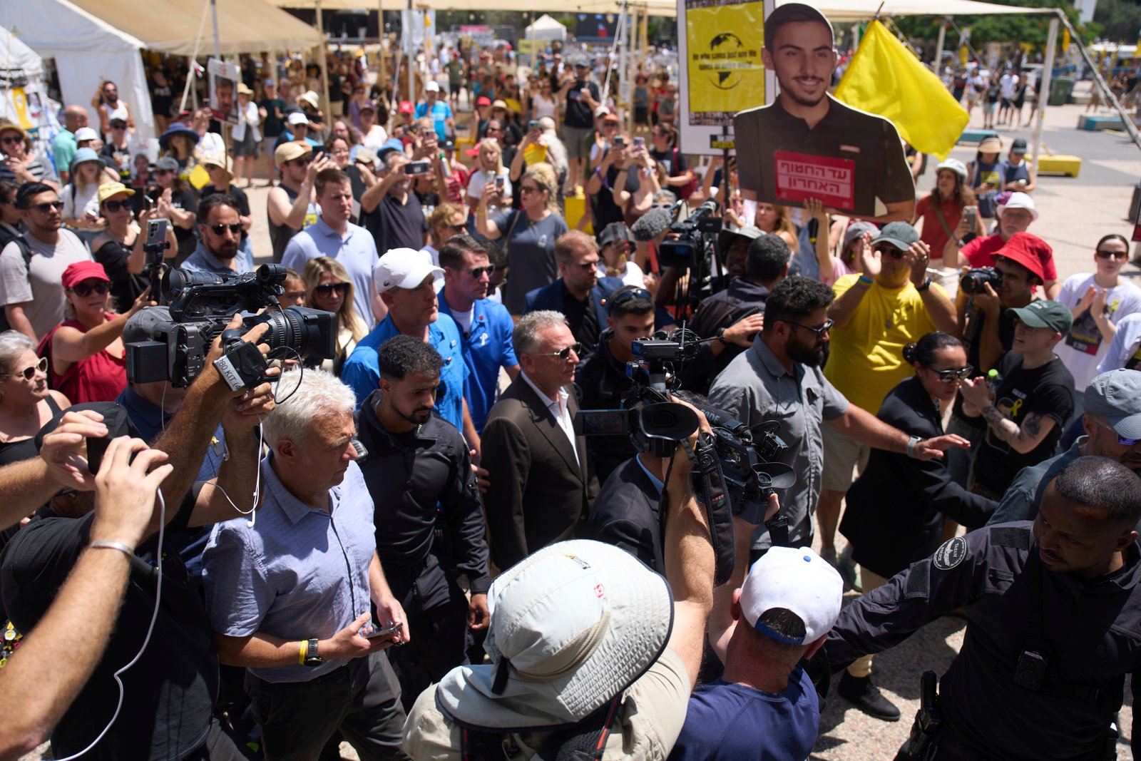 A crowd of people surround a group of people, some of whom are being escorted by security.